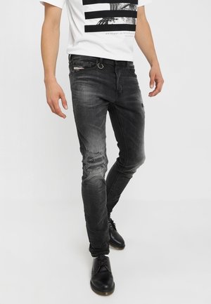 Mann trägt schwarze Slim-Fit-Jeans mit Used-Look, weißes T-Shirt mit Grafik und schwarze Schnürschuhe, steht vor weißem Hintergrund.