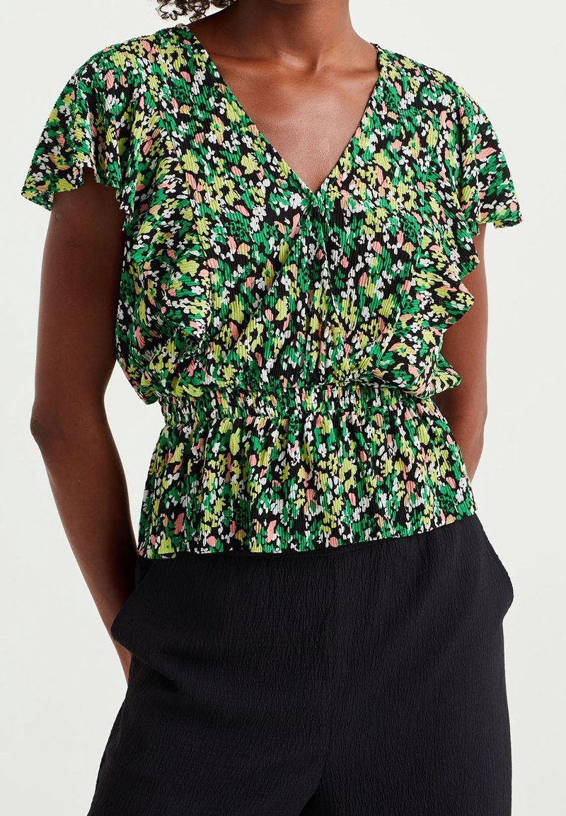 Femme portant un chemisier à volants à fleurs vert, noir et rose avec manches courtes et pantalon noir texturé.