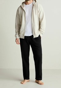 Světlé béžové mikina na zip s texturovaným povrchem, nošená přes bílé tričko a doplněná černými tepláky. Bez viditelných vzorů nebo akcentů.