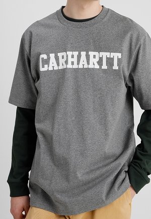 Personne portant un T-shirt gris Carhartt à manches courtes par-dessus une chemise noire à manches longues et un pantalon beige, mains dans les poches.