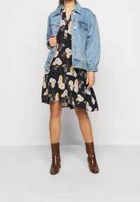 Vrouw in een zwarte bloemenjurk, lichtblauwe spijkerjack en bruine haklaarsjes die tegen een effen achtergrond staat.