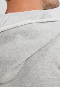 Grå hættetrøje med en glat tekstur. Har snøre i kontrastfarve på hætten og en afslappet pasform. Nærbillede af bagsiden.