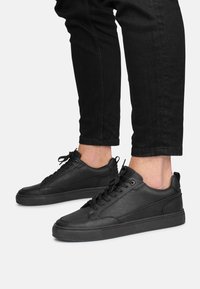 Zapatillas de cuero negro con acabado texturizado, punta redonda, diseño de cordones y suela de goma negra minimalista. Combinadas con pantalones ajustados negros.