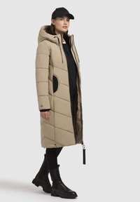 Beige pufferjakke med hætte, med quilted polstring, sidelommer og lynlås lukning. Båret over en sort top og parret med sorte støvler.