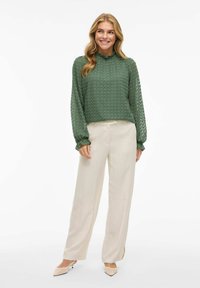 Groene, gestructureerde blouse met lange mouwen en hoge hals, gecombineerd met beige, wijde broekspijpen en lichtgekleurde pumps.