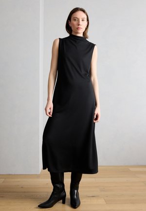 Eine Frau steht drinnen auf einem Holzboden vor einer schlichten hellen Wand und trägt ein ärmelloses schwarzes Midikleid sowie schwarze Stiefel mit Absatz.