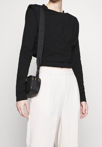 Schwarzes Langarmshirt mit gedrehter Design, kombiniert mit weißen hoch taillierten Hosen. Kleine schwarze Umhängetasche mit goldenen Reißverschlussakzenten.