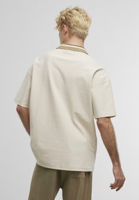 Beige T-Shirt mit kurzen Ärmeln und entspannter Passform, ausgestattet mit einem strukturierten Kragen. Der Stoff wirkt weich und hat eine glatte Oberfläche.