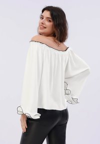Hvit av-skulder bluse med svart trim, lange klokkeermer og volanger på mansjettene. Lett stoff, løs passform.