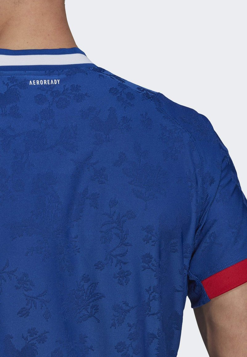 Achteraanzicht van een persoon in een blauwe sportshirt met subtiele bloemmotieven, een wit-blauwe kraag en een rode mouwboord.