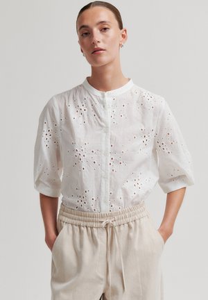 Vrouw draagt een witte blouse met bloemmotief en oogjes, driekwart mouwen, en beige broek met trekkoord, staat met handen in de zakken.