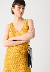 Gul lacedress med V-halsdesign og blomster mønster. Kombinert med en svart håndveske med kjede stropp. Glatt, strukturert stoff.
