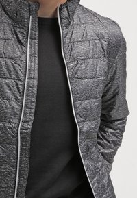 Quiltet grå jakke med en høj krave, struktureret stof og sølvlynlås; lagt over en sort crew-neck sweater.