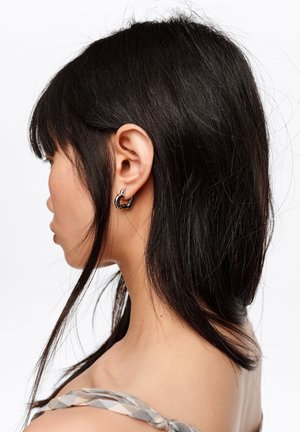 Mujer con cabello liso y negro, lleva pendientes de aro negros y plateados, mirando hacia la izquierda contra un fondo blanco.