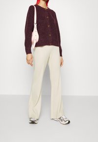 Burgundy, fluffig stickad kofta med guldknappar, kombinerad med krämfärgade ribbade utsvängda byxor och metalliska sneakers. Ljusrosa axelväska.