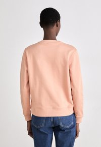 Sudadera color melocotón hecha de material suave, con un escote redondo, mangas largas y un dobladillo acanalado. Llevada con jeans de mezclilla azules.