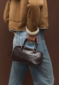 Braune Lederhandtasche mit rechteckiger Form, doppelten Tragegriffen und goldfarbenen Beschlägen, kombiniert mit hellblauem Denim und einer braunen Jacke.