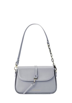 Femme portant un haut noir sans manches et un pantalon foncé, tenant un petit sac blanc à bandoulière avec une boucle sur un fond neutre.