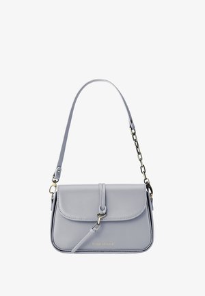 Femme portant un haut noir sans manches et un pantalon foncé, tenant un petit sac blanc à bandoulière avec une boucle sur un fond neutre.
