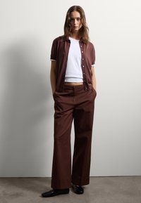 Camisa marrón de manga corta y botones superpuesta sobre una camiseta blanca corta, combinada con pantalones marrones de pierna ancha y zapatos negros.