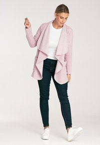 Jasnoróżowy dziergany kardigan z przodem w stylu drapowanym, bocznymi kieszeniami i długimi rękawami, zestawiony z ciemnymi dżinsami i białymi trampkami.