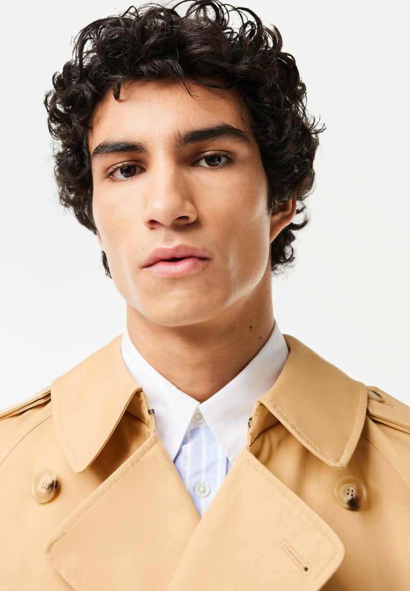 Trench-coat beige avec un design à double boutonnage, de grands boutons et un large col, porté sur une chemise blanche à rayures. Texture de tissu lisse.