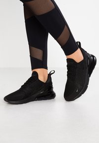 Ben i svarta leggings med meshpaneler, iförda svarta Nike Air Max 270-sneakers på ett vitt golv med ena foten något höjd.