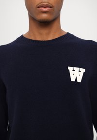 Pulover din lână bleumarin, cu gât rotund și margine ribată, având un logo alb "W" proeminent pe piept. Textură netedă.