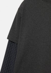 Haut à manches courtes gris foncé superposé à une chemise à manches longues à carreaux noirs et gris, visible de l'épaule jusqu'au milieu du torse.