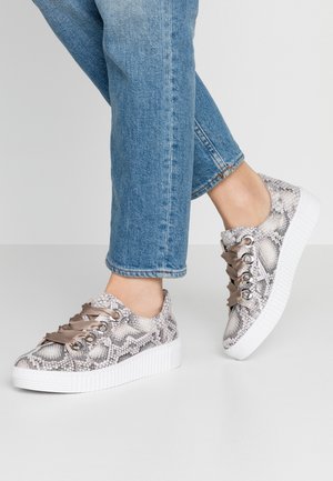 Fødder iført blå jeans og platformsneakers med slangeskinsmønster og brede satin snørebånd på en lysegrå gulv.