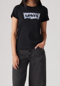 Kvinna iklädd svart Levi's t-shirt med blå mönstrad logotyp och lösa mörkgrå jeans, stående med en hand i fickan mot en enfärgad bakgrund.