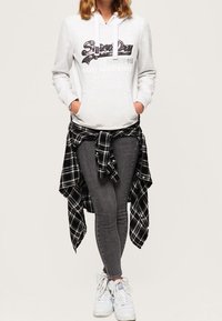 Felpa grigia con zip e tasca frontale, scritta nera. Camicia a scacchi bianca e nera legata in vita, jeans grigi attillati, sneakers bianche.