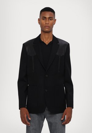 Blazer negro de tejido texturizado, con acentos de cuero en los hombros, dos bolsillos frontales y un cierre de un botón. Diseño de corte recto.
