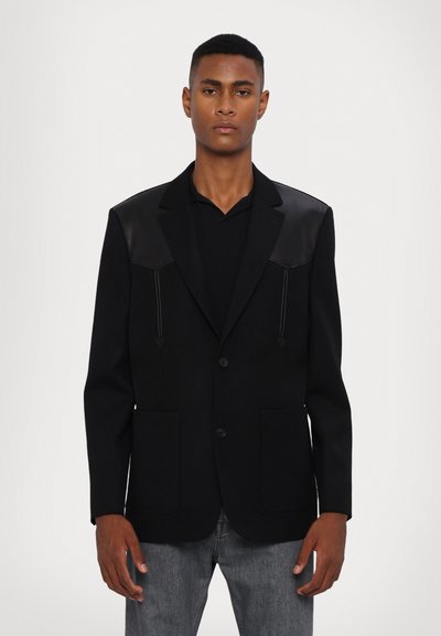 Blazer noir en tissu texturé, avec des accents en cuir aux épaules, deux poches avant et une fermeture à un bouton. Design à coupe droite.