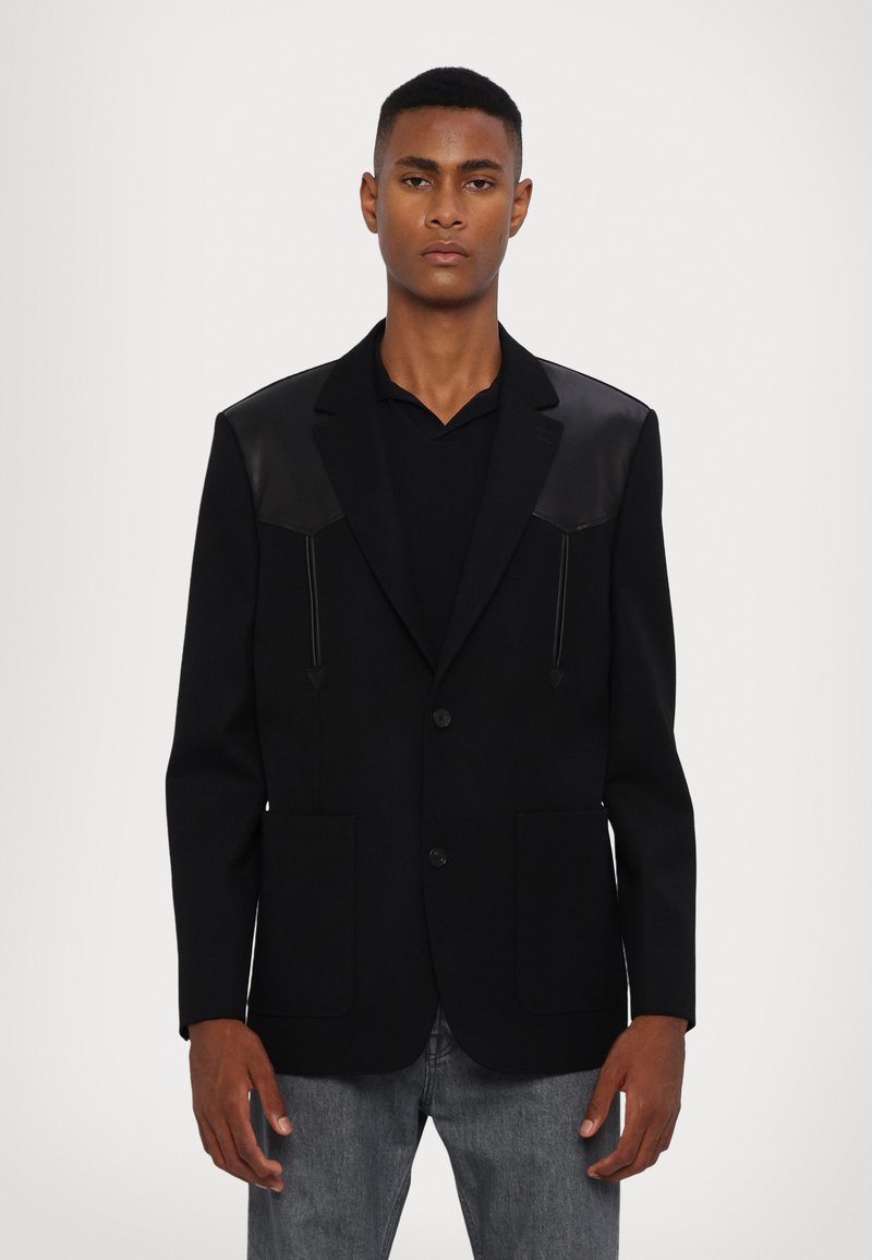 Zwarte blazer van gestructureerde stof, met leren schouderaccenten, twee voorzakken en een enkele knoopsluiting. Recht model.