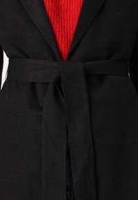 Blazer noir à ceinture avec un tissu texturé, présentant un gilet rouge en tricot visible à l'intérieur. La ceinture est nouée à la taille, mettant en valeur la silhouette.