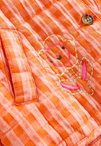 Orange checkered fabric with a small pocket, a brown button, and a stitched smiling bear holding pink leaves.
