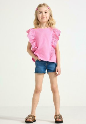 Lyshåret pige står med hænderne ned langs siden iført en pink flæset top, denimshorts, sandaler med leopardprint og pink hårspænder på hvid baggrund.