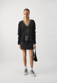 Maglietta nera a maniche lunghe con scollo a V e chiusura con lacci, decorata con ricami a motivo foglia beige. Abbinata a una gonna di jeans nera e scarpe da ginnastica grigie.
