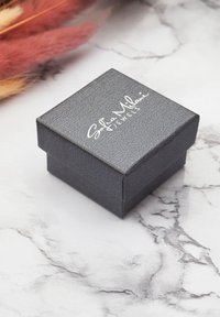 Svart texturerad smyckeslåda med lock, med vit präglad varumärkeslogotyp. Fyrkantig form, minimalistisk design, mot en marmorbakgrund.