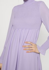 Levandulové šaty se smockovaným živůtkem, vysokým výstřihem a dlouhými rukávy. Znaky plynoucí sukně a texturované manžety pro přidaný detail.