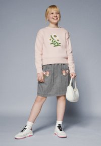 Rosa strikket genser med blomsterbroderi, grå skjørt med hjerteformede lommer, hvite og rosa sneakers, holder en liten hvit håndveske.