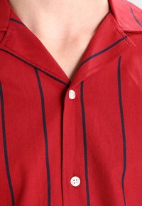 Rode shirt met een camp collar, voorzien van smalle marineblauwe verticale strepen en een knoopsluiting. De stof lijkt textuur te hebben en is geweven.