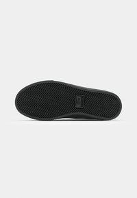 Suola di sneaker nera con un motivo a lisca di pesce e un logo rettangolare al centro. Il materiale sembra gomma con superficie di presa testurizzata.