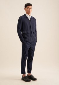Giacca camicia in lino blu navy con due tasche frontali, abbinata a pantaloni su misura dello stesso colore. Modello che indossa scarpe in pelle nere.