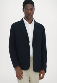 Navy blazer with two buttons and notch lapels, made from textured fabric. Worn over a white shirt, paired with light beige trousers.