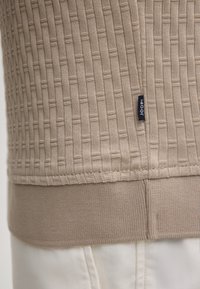 Orlo beige a maglia strutturata di un maglione con polsino a coste e piccolo etichetta in tessuto nero con la scritta "JOOP!" sopra pantaloni bianchi.