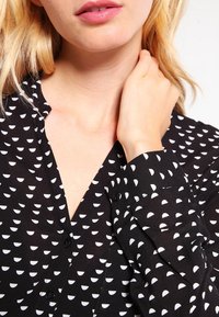 Czarna bluzka zapinana na guziki z półokrągłym białym wzorem, długimi rękawami z mankietami na guziki oraz z kołnierzykiem. Miękki, lekki materiał.