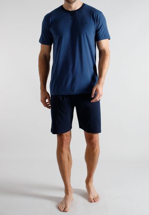 Mann steht barfuß und trägt ein blaues Kurzarmhemd sowie marineblaue Shorts vor einem schlichten hellen Hintergrund.