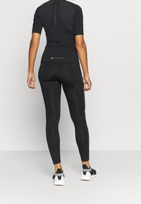 Osoba ubrana w czarne legginsy sportowe, dopasowaną koszulkę z krótkim rękawem oraz czarno-białe sneakersy, stojąca na tle jednolitym.